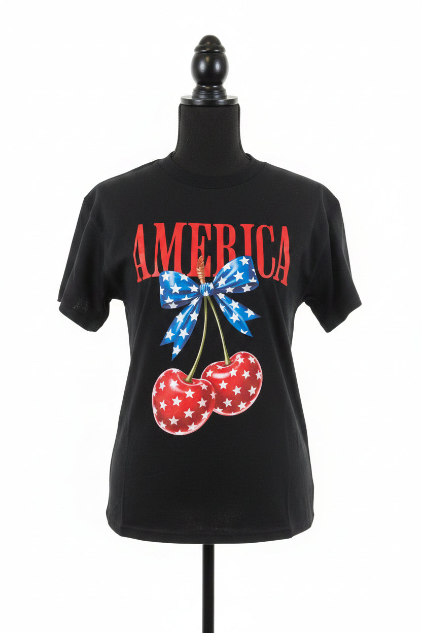 Black t-shirt with 'AMERICA' text, bow, and cherries design on a red brick wall background