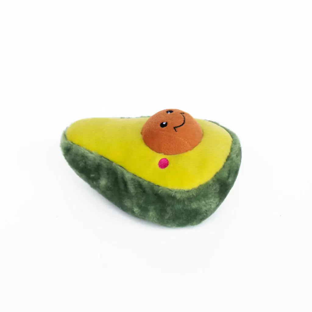 A plush avocado toy with a green outer skin, yellow-green inner flesh, a brown smiling seed, and a small pink accent, photographed on a plain white background.