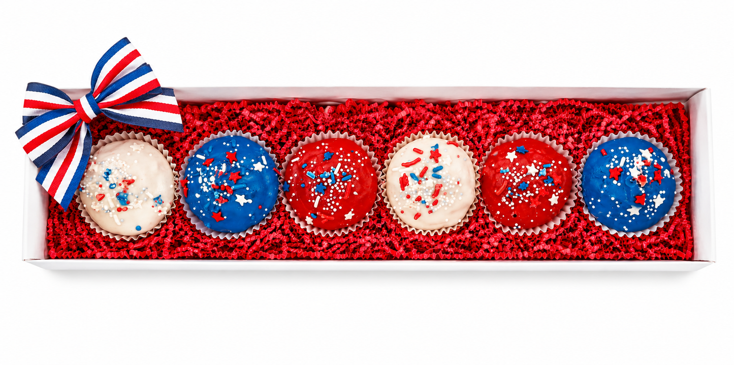 Six patriotic-themed cupcakes decorated in red, white, and blue, presented in a white gift box filled with red crinkle paper and topped with a matching red, white, and blue bow.