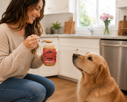 A woman in a beige sweater and blue jeans kneels in a bright kitchen, holding a jar of 'Buddy Budder' raw bacon spread and offering a spoon to a golden retriever dog. The dog looks up at the jar with interest.