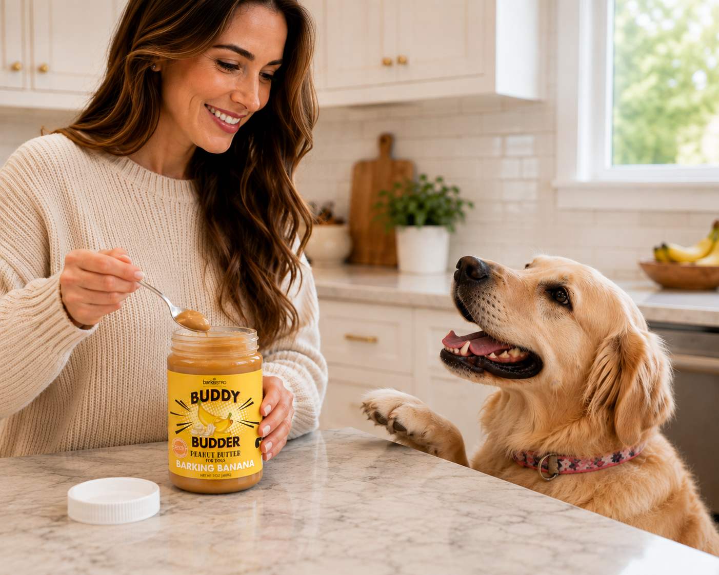 A woman in a cream knit sweater smiles while offering peanut butter from a jar labeled 'Buddy Budder Peanut Butter' to a golden retriever dog standing at a kitchen counter.