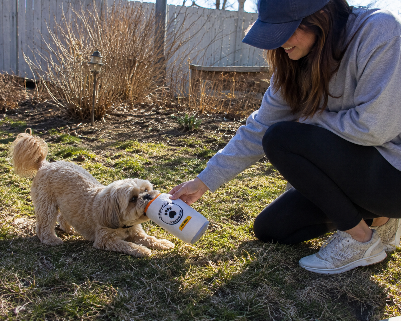 A woman crouches outdoors on grass while holding a white dog toy bottle with a black paw print logo, offering it to a small cream-colored dog.